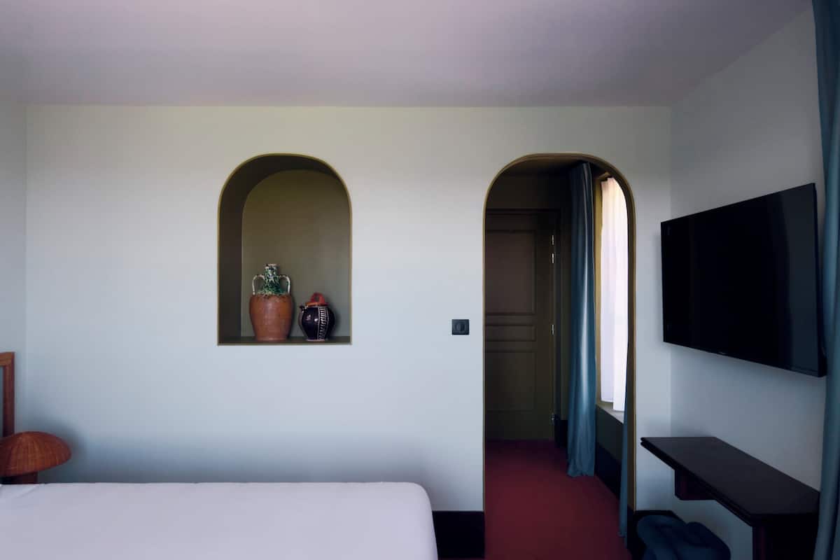 Habitación de hotel con cama, televisor montado en la pared, una ventana pequeña con vistas y una puerta que da a otra habitación.