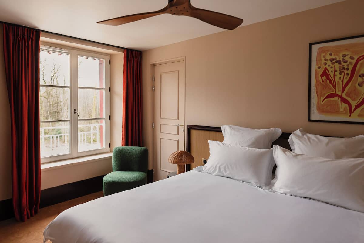 Una habitación de hotel con cama, una silla, ventilador de techo, ventana con cortinas y una obra de arte enmarcada en la pared.