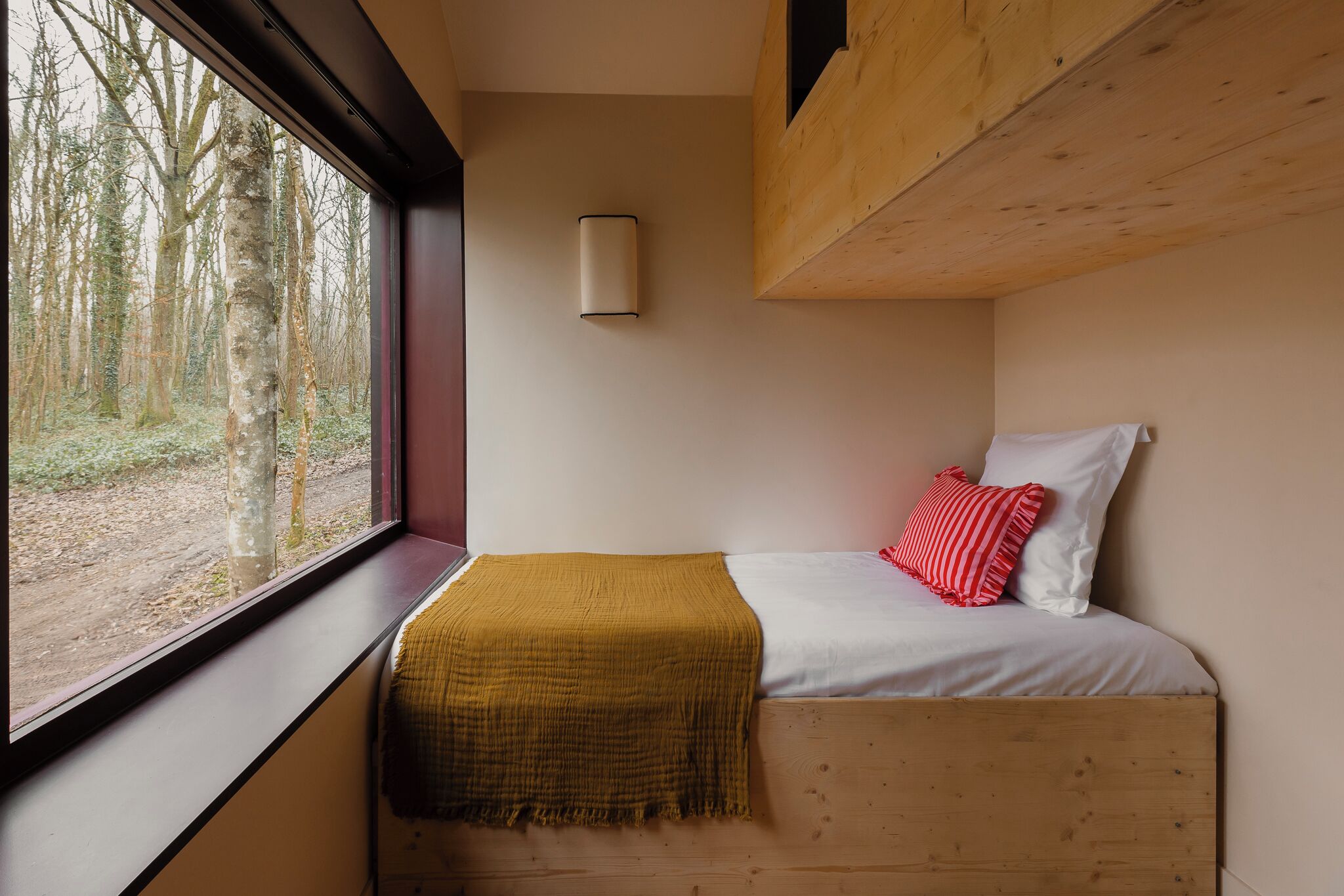 A small, cozy bedroom with a window overlooking a forest, a bed with white and red bedding, and a mustard-colored throw blanket.