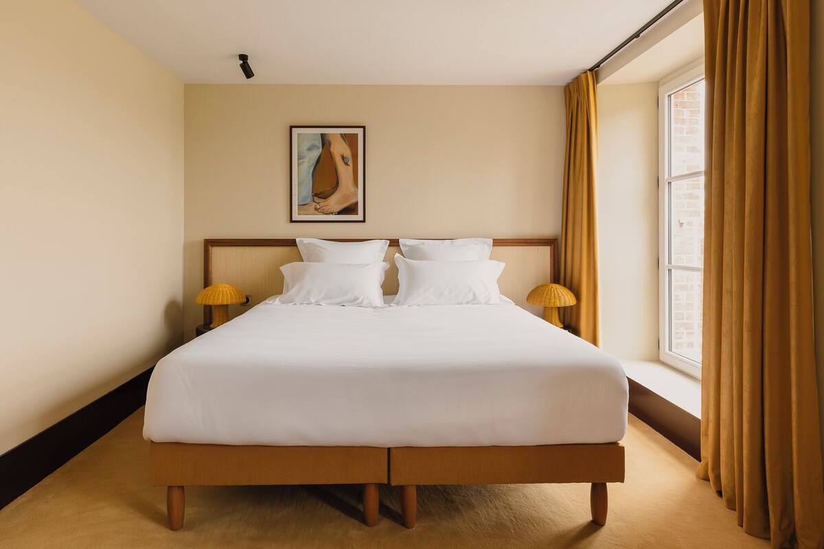 Una cama bien hecha con sábanas blancas, dos lámparas de noche, una obra de arte enmarcada en la pared y una ventana con cortinas.