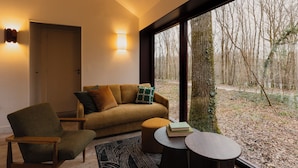 A modern living room with a sofa, armchair, and a small round table. A door and a window with a view of a forest are visible. - Repère Sauvage, Hôtel Lodges & Restaurant (La Ville-aux-Clercs)