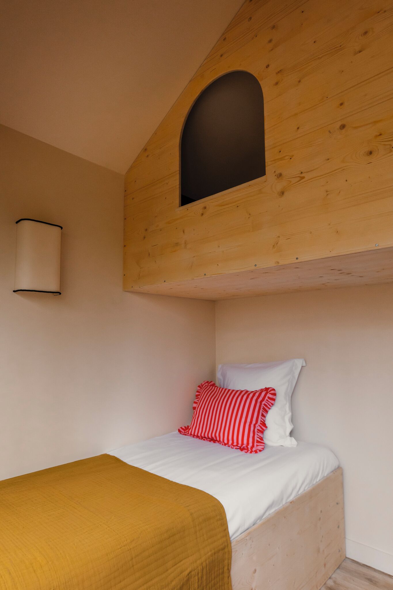 Un lit simple avec une tête de lit en bois, une petite fenêtre et un luminaire fixé au mur.