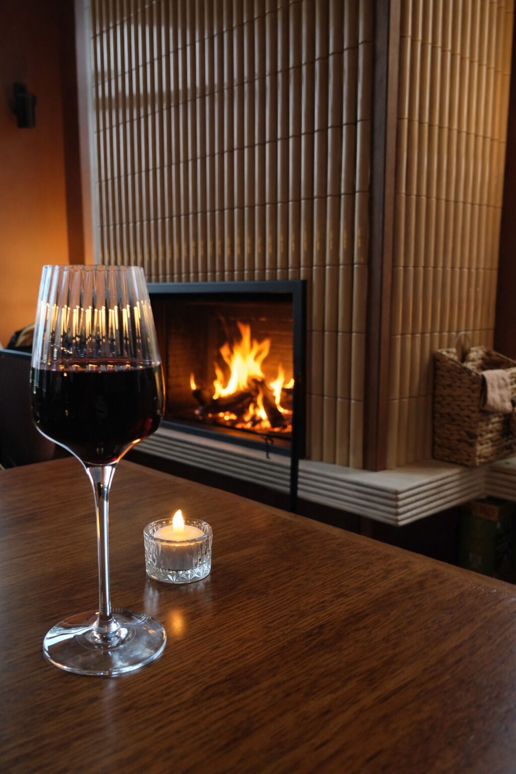 A wooden table with a glass of red wine and a lit candle, in front of a fireplace with a visible fire.