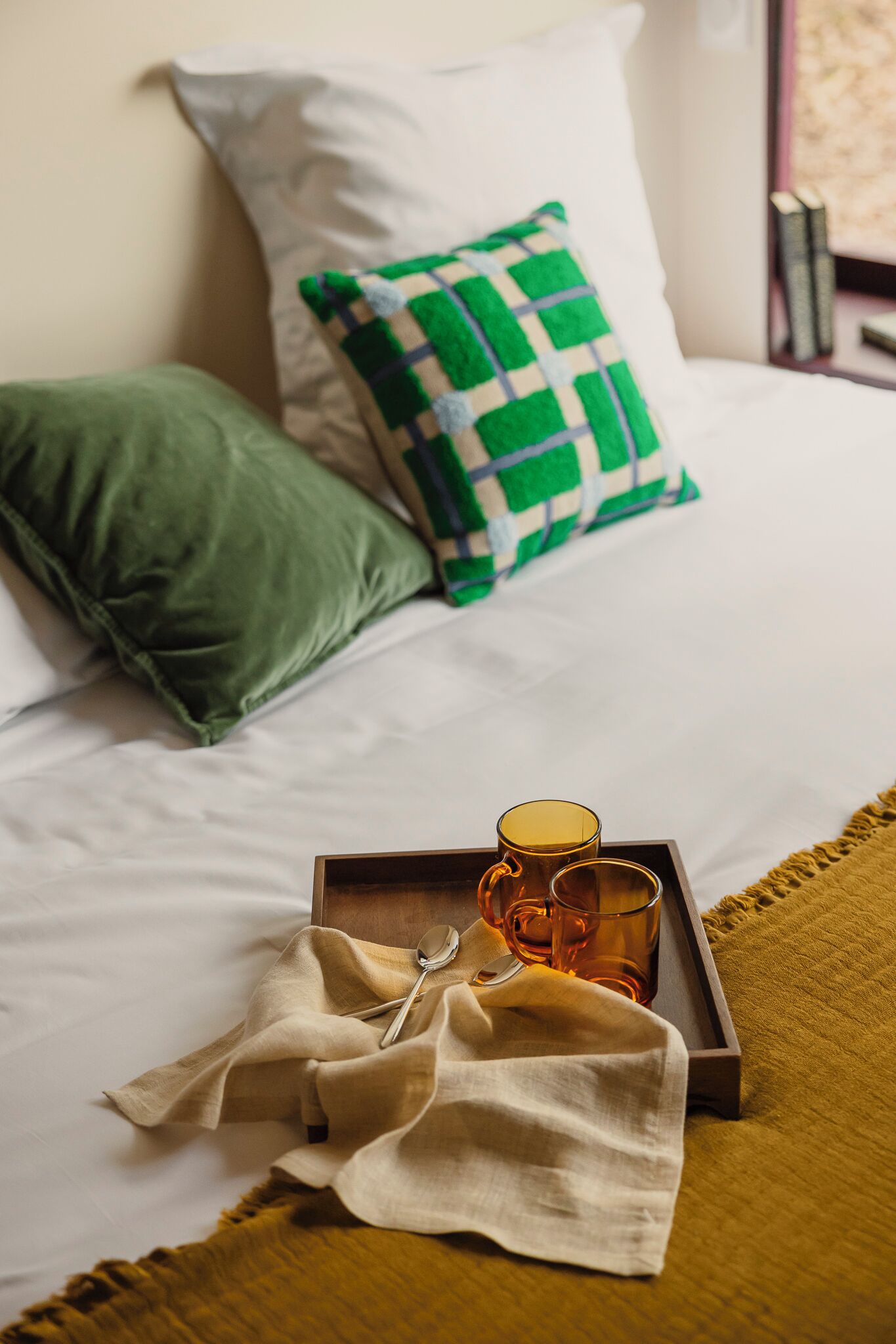 A neatly made bed with white sheets, a green and white checkered pillow, and a green pillow. A tray with a mug and a spoon is placed on the bed.
