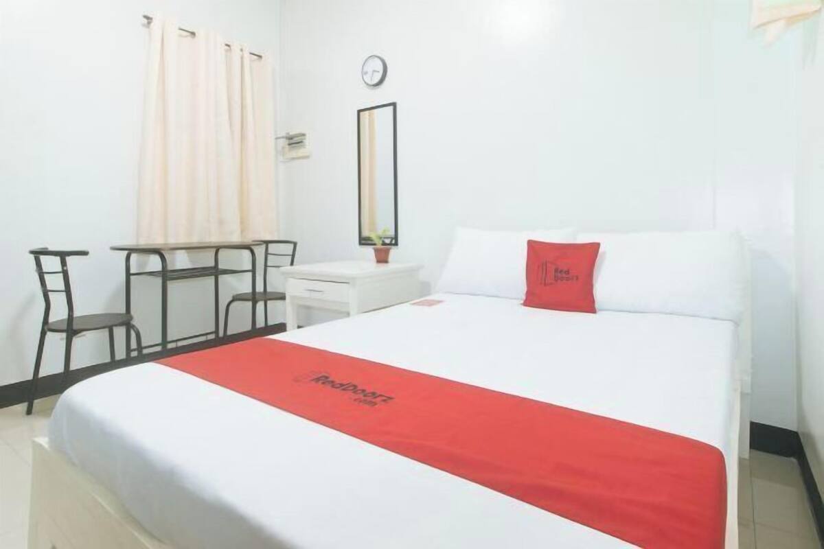 Habitación de hotel con cama, escritorio, silla, una mesita, un reloj en la pared y una ventana con cortinas.