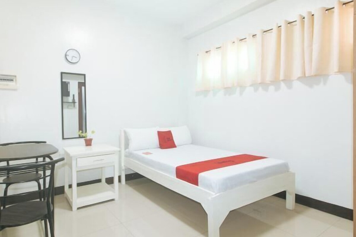 Une chambre avec un lit simple, un petit chevet, une chaise, un miroir et une horloge au mur.