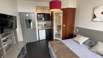 Une chambre d’hôtel de taille réduite, comprenant un lit, un bureau, une kitchenette et une salle de bain.