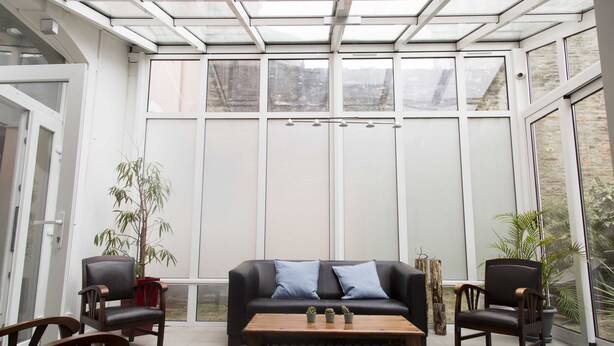 A conservatory with a glass ceiling, a black leather sofa with blue cushions, two wooden chairs, and a wooden coffee table with small plants. - Smart Appart Cherbourg La Rose des vents (Cherbourg-en-Cotentin)