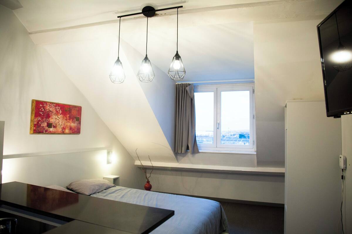 A bedroom with a bed, bedside table, lamp, window, and artwork.