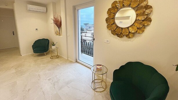 Un salon moderne avec un fauteuil vert, un miroir rond doté d’un cadre en forme de feuille, et une vue à travers une porte vitrée.