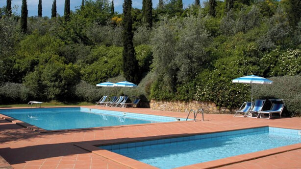 Una piscina rectangular con azulejos azules, rodeada por una terraza de ladrillos rojos. Hay sillas de descanso azules y sombrillas a lo largo de la piscina.