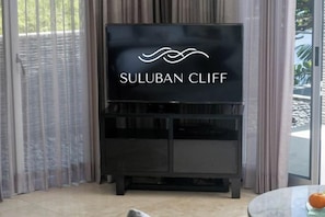 A black entertainment unit with a flat-screen TV displaying 'SULUBAN CLIFF' and a small table with a glass top and an orange fruit. - Suluban Cliff Bali Villa (Bali)