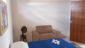 Una habitación con un sillón color beige, una mesita con un ventilador y un mantel azul con diseño de flores blancas.