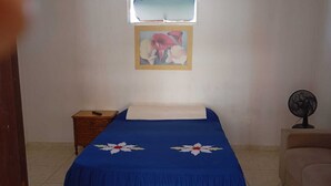 Un dormitorio con cama, mesita de noche, silla, ventilador de pared y un cuadro.