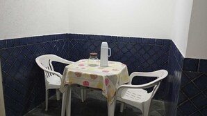 Un pequeño comedor cerrado con una mesa redonda cubierta por un mantel floral, dos sillas de plástico blancas y una jarrón blanco sobre la mesa.