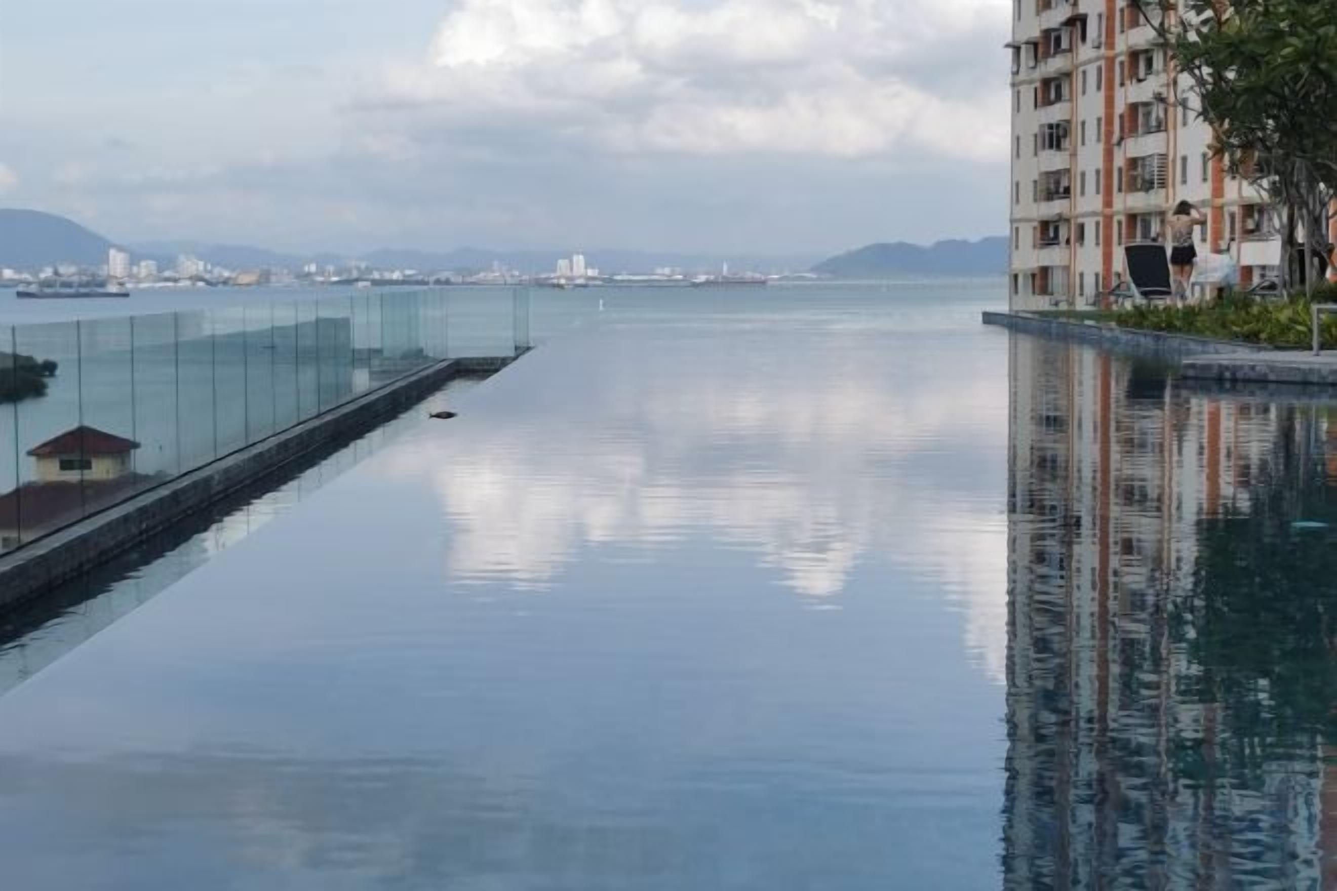 A long, narrow swimming pool with a glass barrier on one side, a view of a distant cityscape, and a building with balconies on the other.