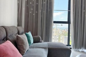 Ein modernes Wohnzimmer mit einem grauen Sofa, zahlreichen Kissen und einem großen Fenster mit Blick auf die Stadt.