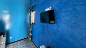 A room with blue walls, a window with patterned curtains, a small shelf with items, and a trash bin labeled "NEW". - UBANGUI residence. A unique, calm and inspiring setting! Suitable for creation! (Gandon, Saint-Louis)