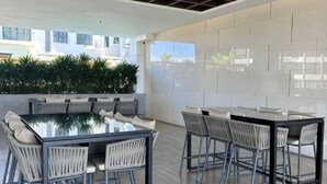 Un área de comedor exterior moderna con una mesa de vidrio y sillas de metal, rodeada por un muro blanco y vistas a edificios.