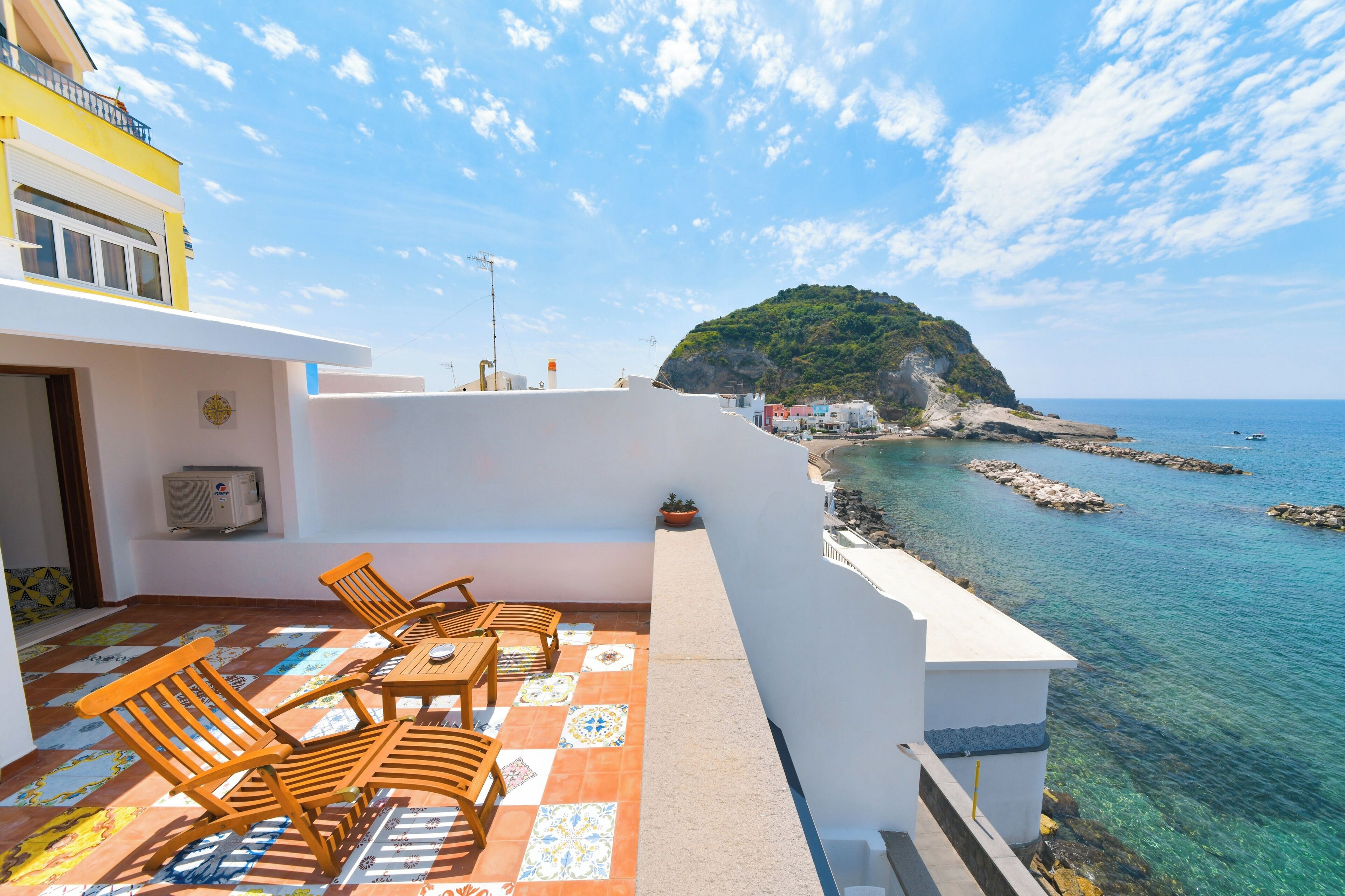 A terrace with a view of the sea, featuring a table and chairs, a potted plant, and a building with a balcony.