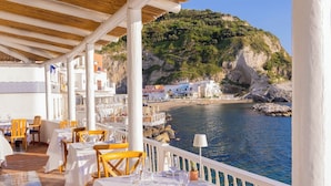 Un restaurant en bord de mer avec une terrasse, des tables blanches et des chaises jaunes, offrant une vue sur une côte rocheuse.