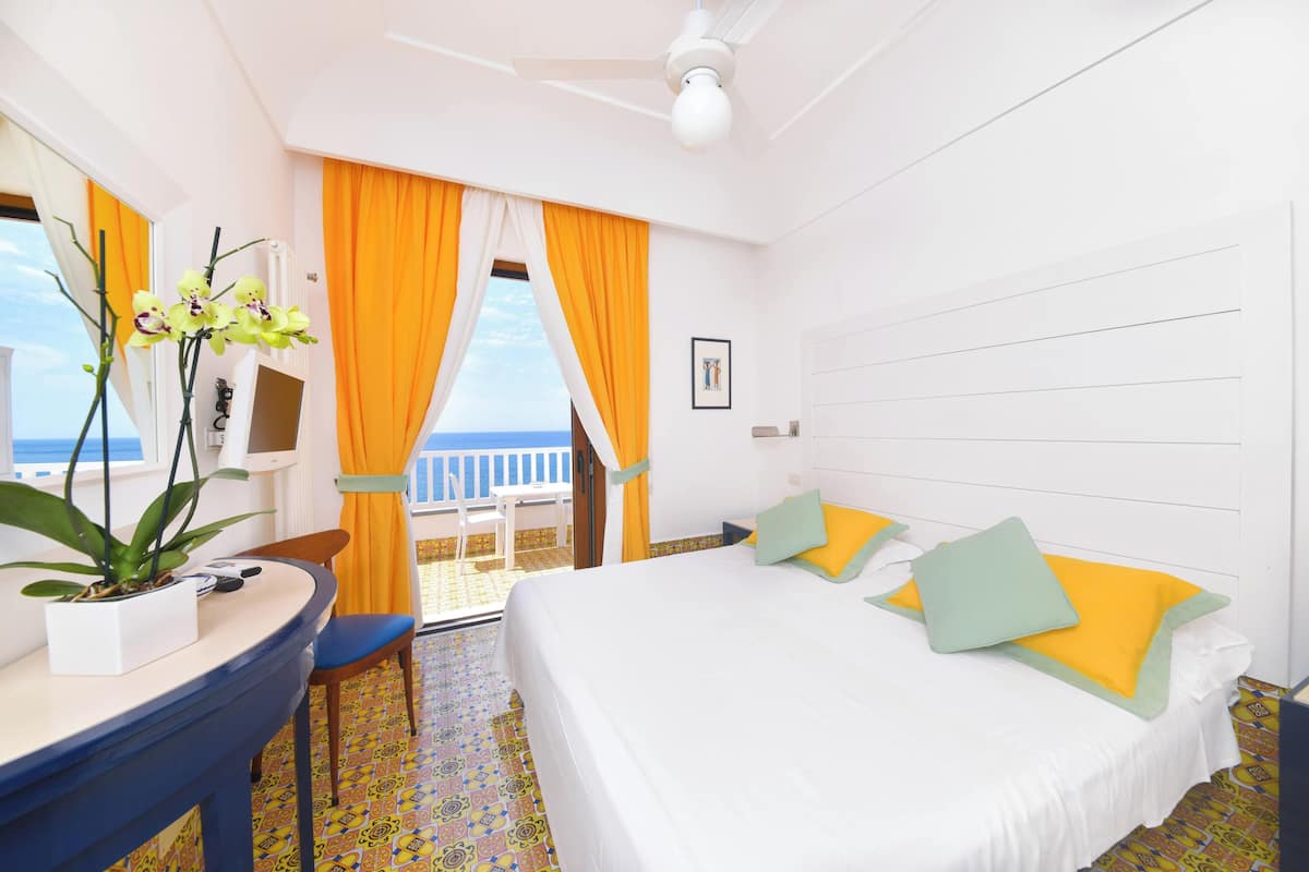 Une chambre d’hôtel avec un lit, un bureau, une chaise, une plante en pot et une vue sur la mer.