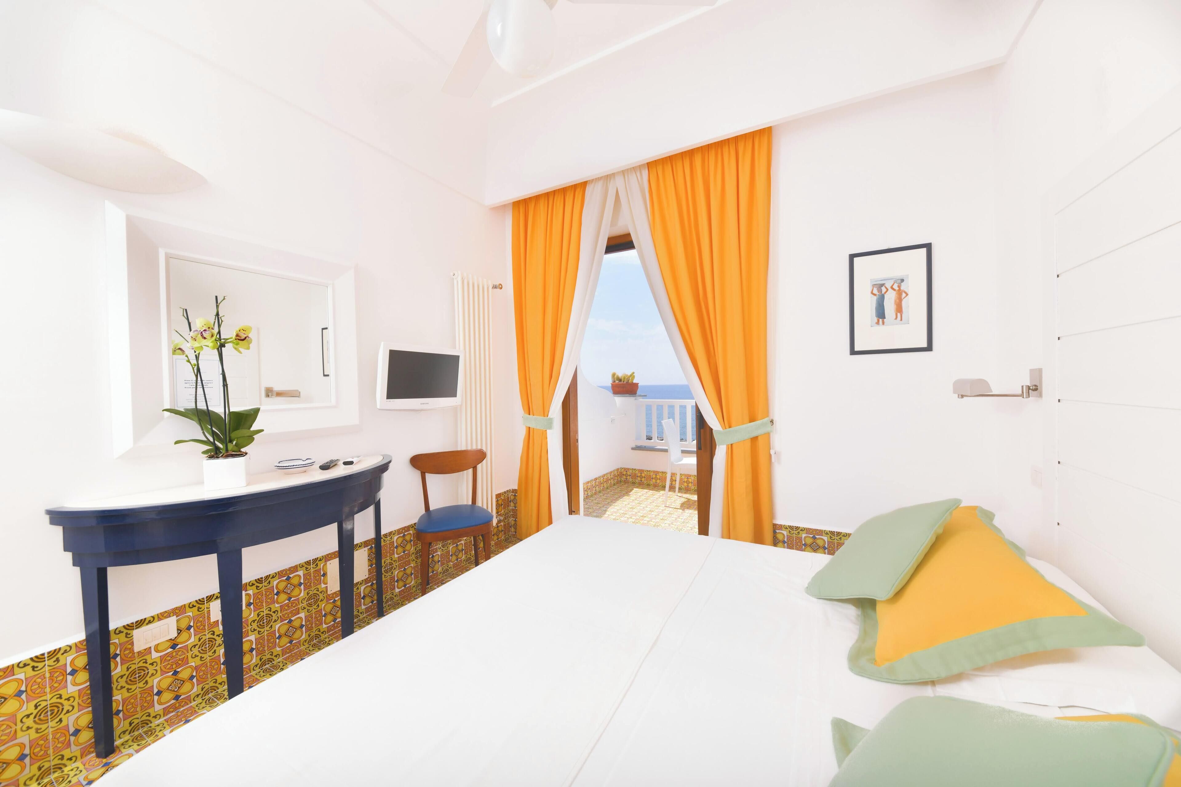 Hotelkamer met een bed, een bureau, een stoel, een plant in pot en uitzicht op zee door gordijnen.