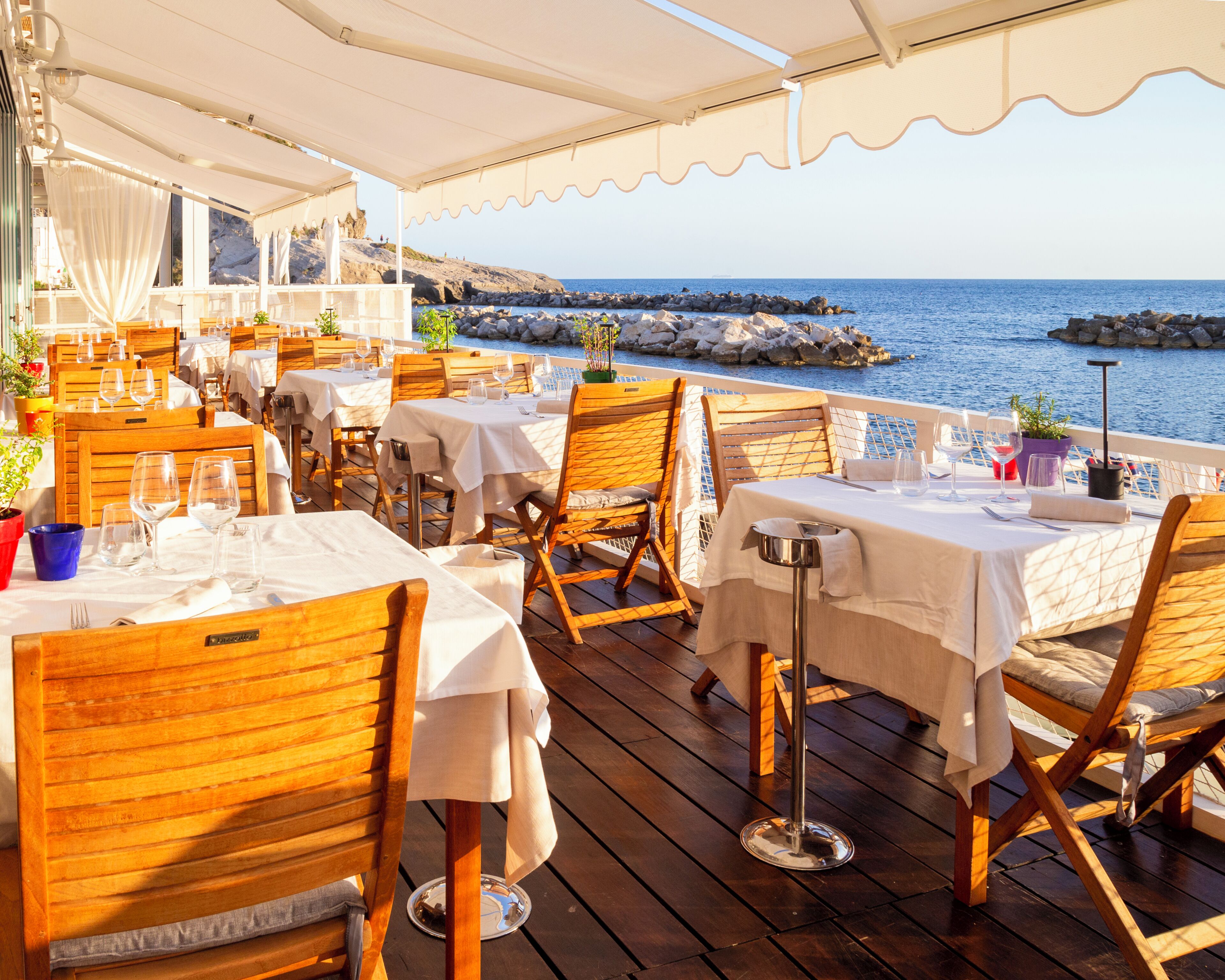 A seaside restaurant with outdoor seating, tables, chairs, and a view of the sea.