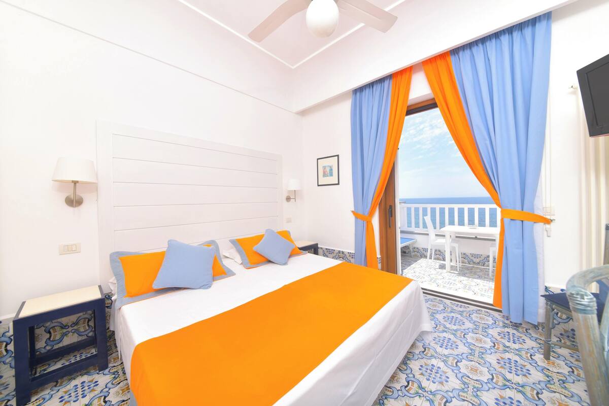 Une chambre d’hôtel avec un lit, une vue sur la mer et un balcon agrémenté de rideaux bleus et oranges.