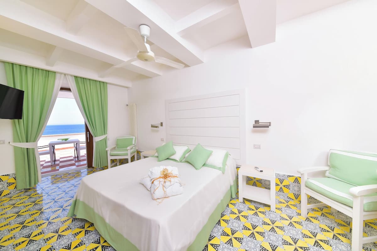 Une chambre d’hôtel avec un lit, des rideaux verts, un ventilateur de plafond, une télévision et une vue sur la mer.