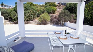 A covered outdoor seating area with a white table and chairs, a blue cushioned bench, and a view of a hillside with trees and buildings. - Almyra Luxury Pool Suite with Private Pool (Πάνορμος)