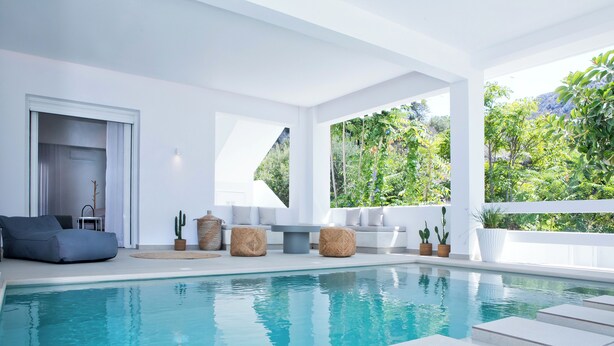 A modern indoor pool area with a clear view of the surrounding greenery and a seating area with cushions and planters. - Almyra Luxury Pool Suite with Private Pool (Πάνορμος)