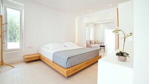 A modern bedroom with a wooden platform bed, a small seating area, and a potted orchid. - Almyra Luxury Pool Suite with Private Pool (Πάνορμος)