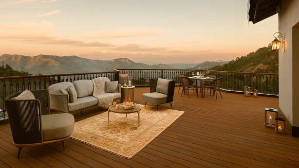 Una terraza de madera con mobiliario exterior, una mesa y vistas a montañas distantes.