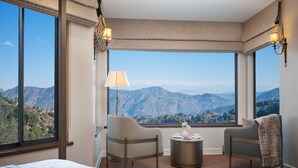 Habitación de hotel con un ventanal amplio que ofrece vistas a la montaña, una cama, dos sillas, una mesita con un florero y dos lámparas de pared.