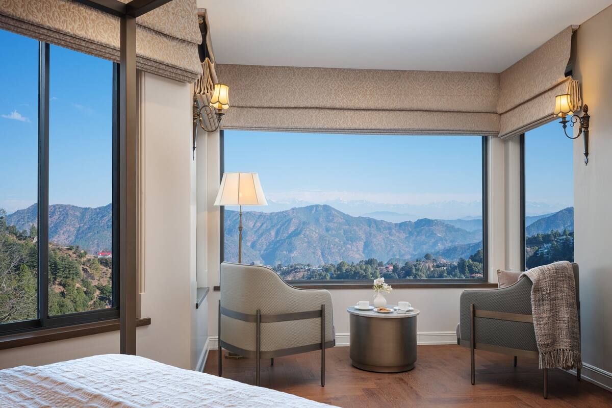 Habitación de hotel con un ventanal amplio que ofrece vistas a la montaña, una cama, dos sillas, una mesita con un florero y dos lámparas de pared.