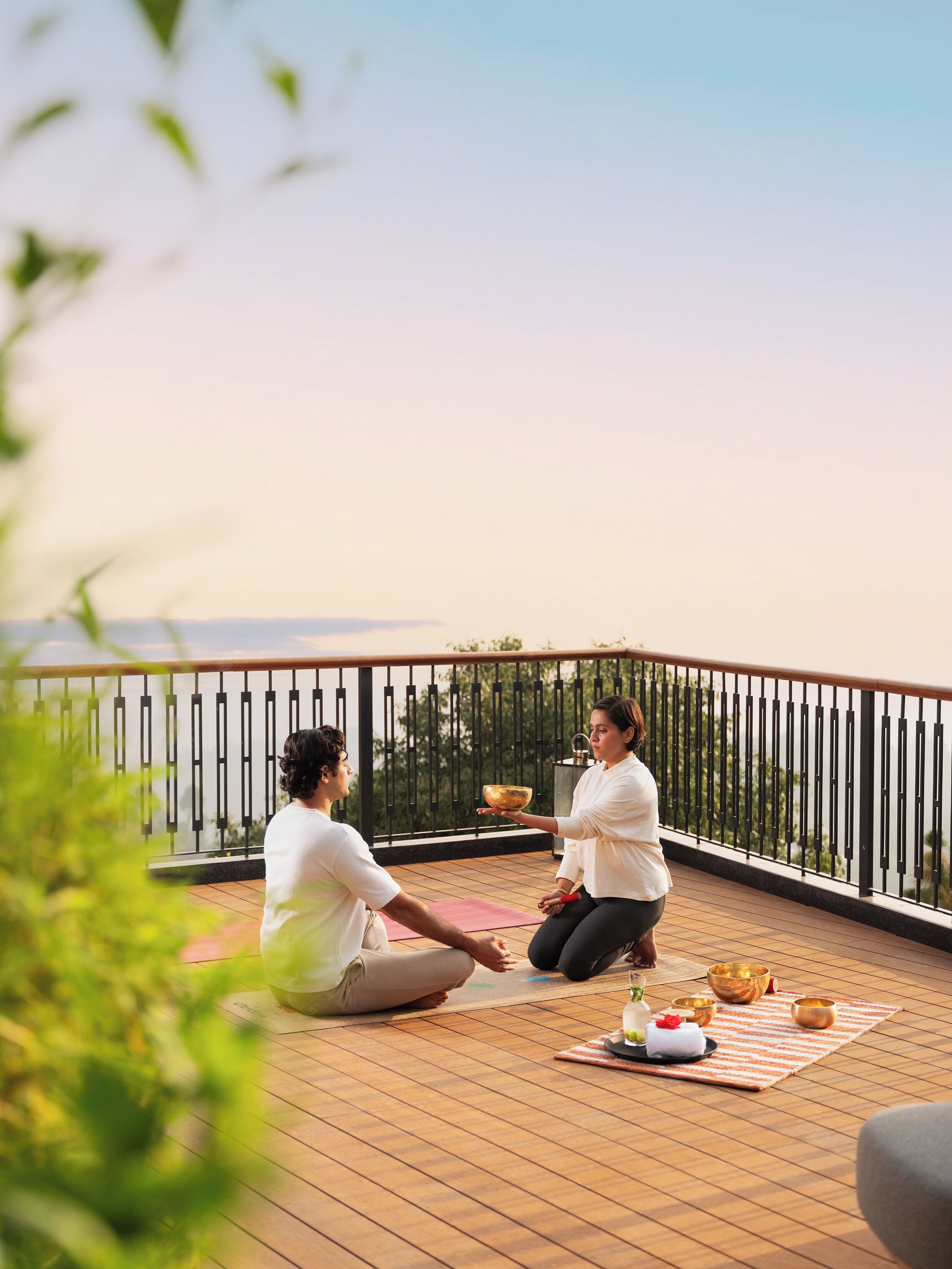 Two people sitting on a wooden deck outdoors, engaged in a meditation session with a bowl and other items on the floor.