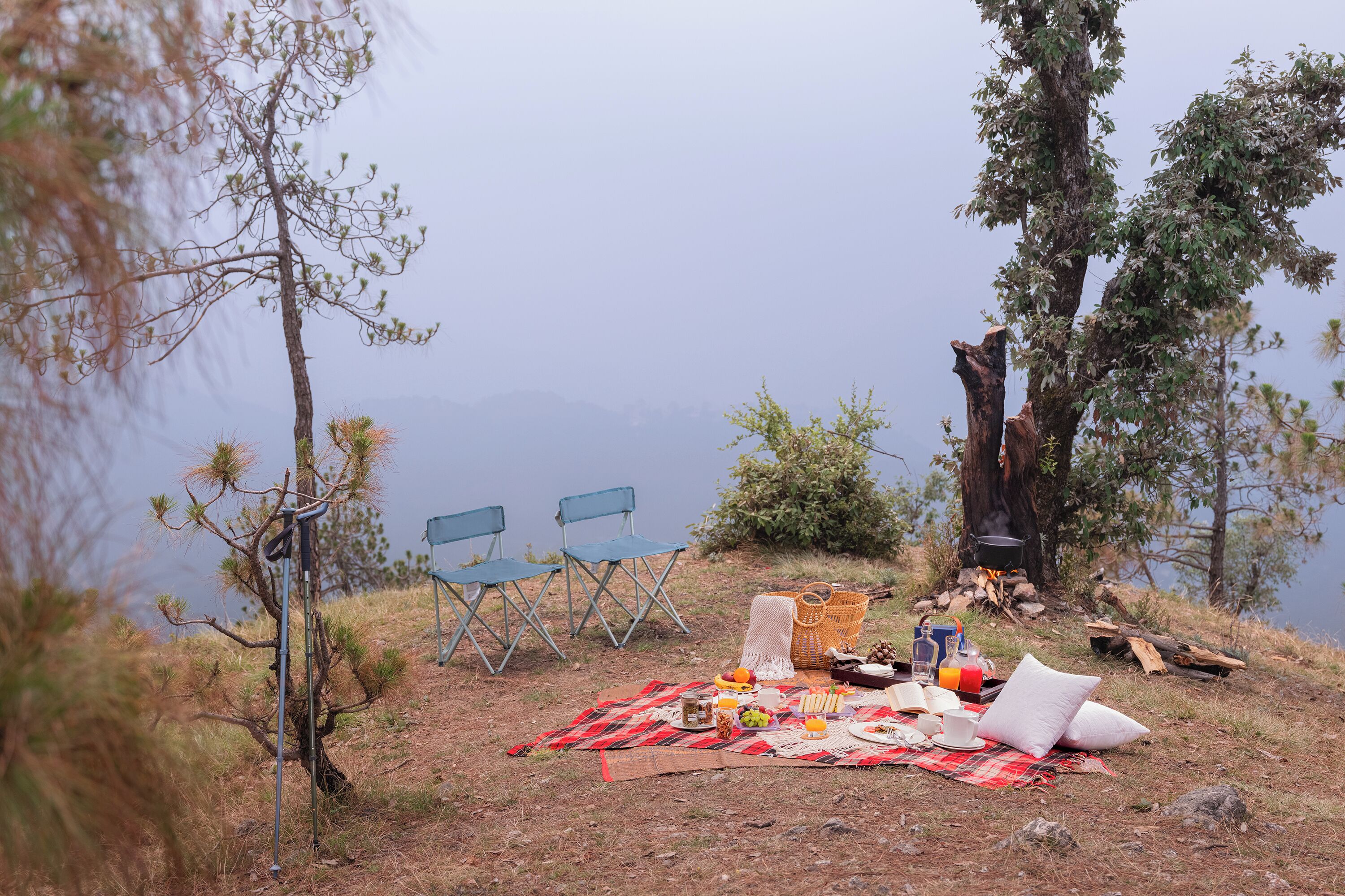 A picnic setup with a blanket, food, and chairs by a lake with mountains in the background.
