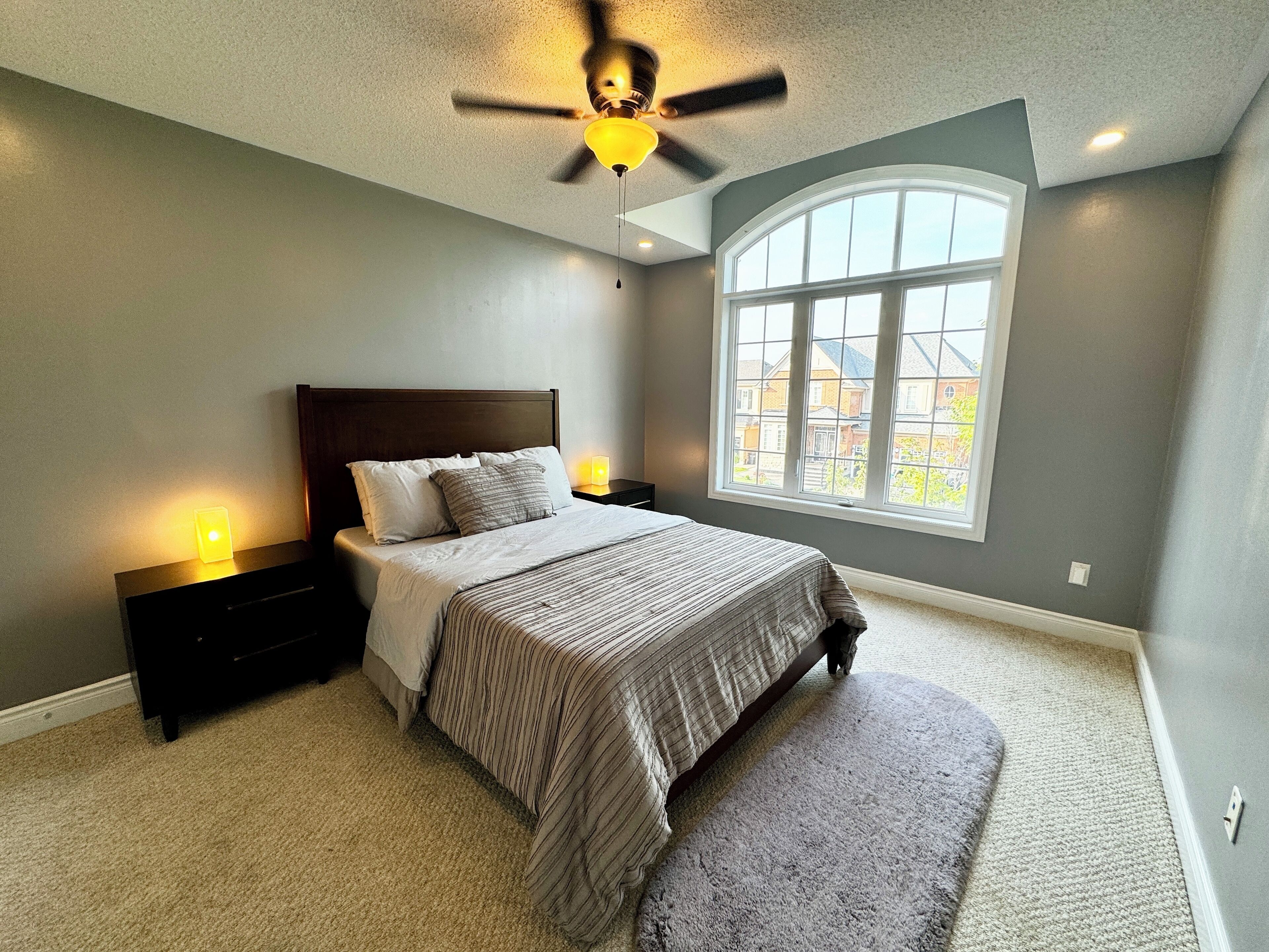 A bedroom with a large bed, a nightstand, a ceiling fan, a window with a view, and a carpeted floor.
