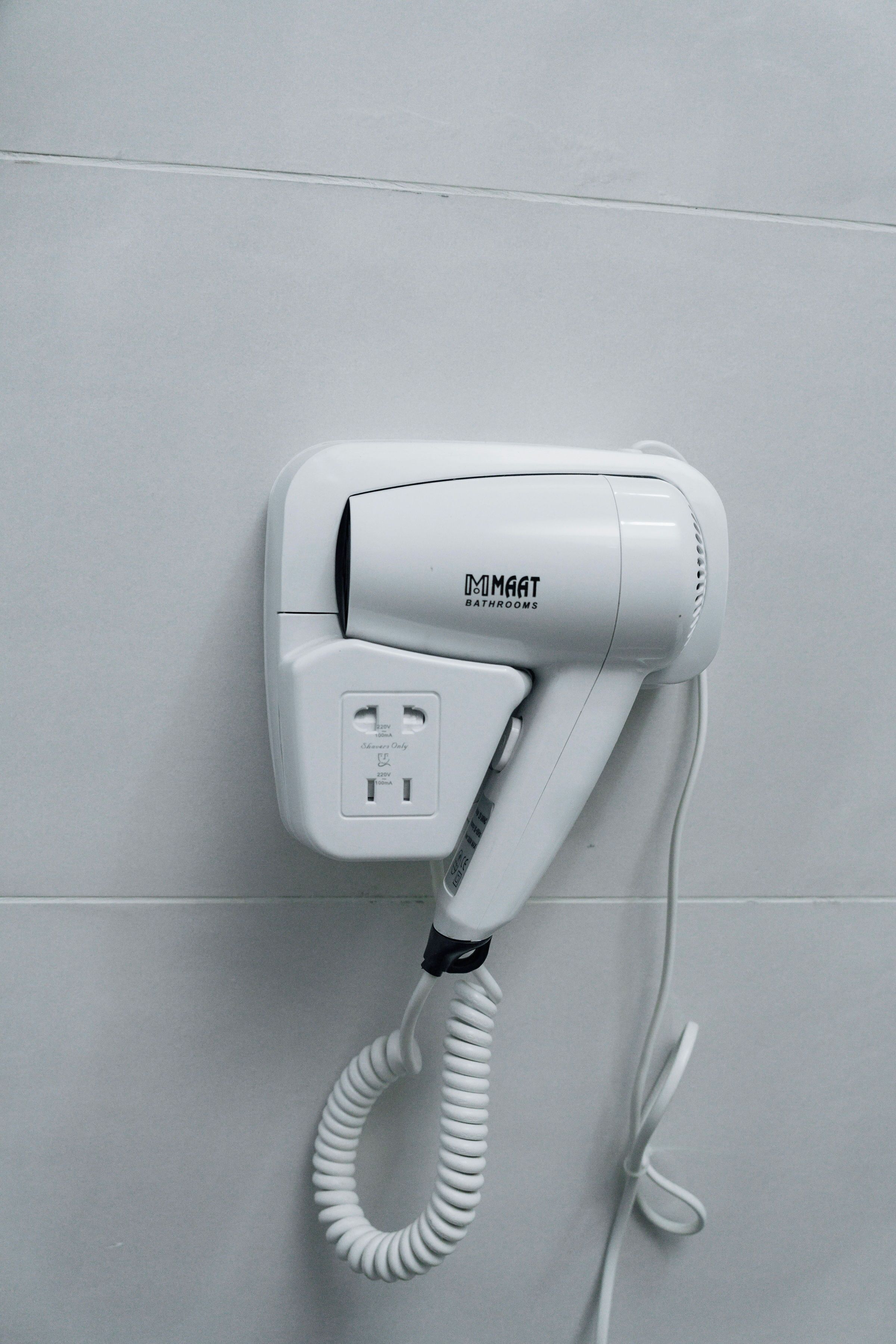 A white hair dryer mounted on a tiled wall, with a coiled cord and a power plug.