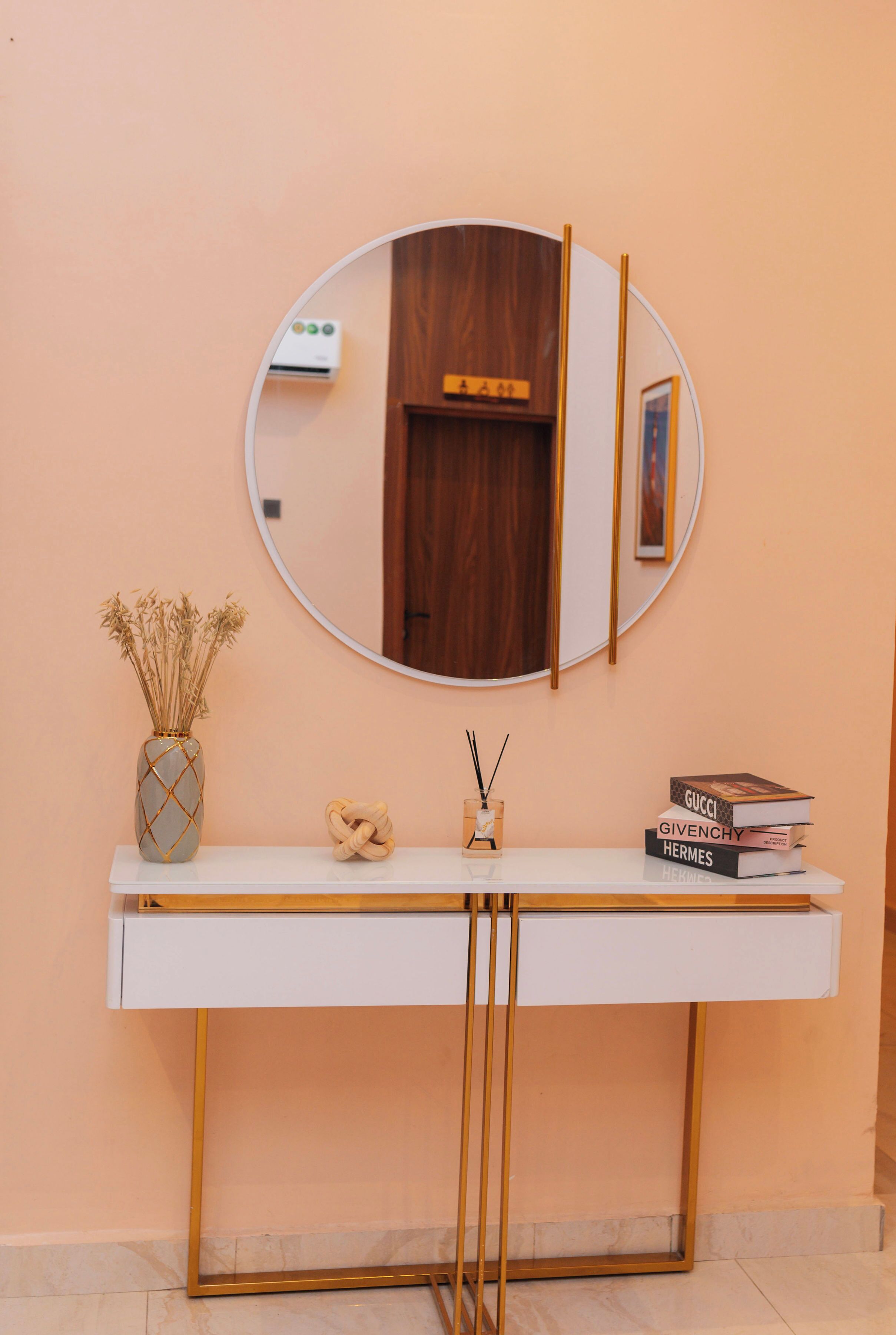 A white console table with a round mirror above it, a vase with dried flowers, a small sculpture, and a stack of books.