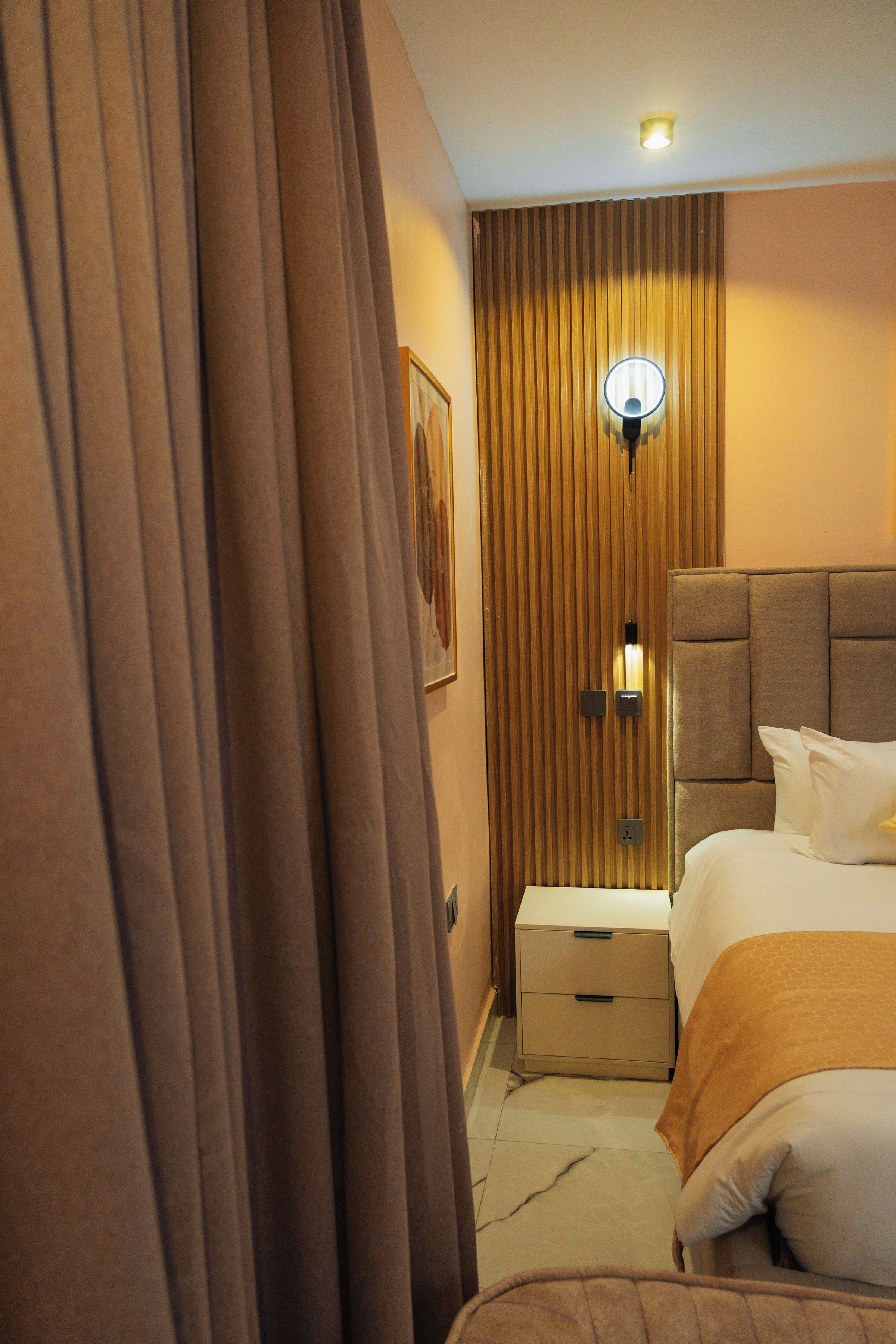 Una habitación de hotel con cama, mesita de noche, cajón, lámpara de pared y cortina.