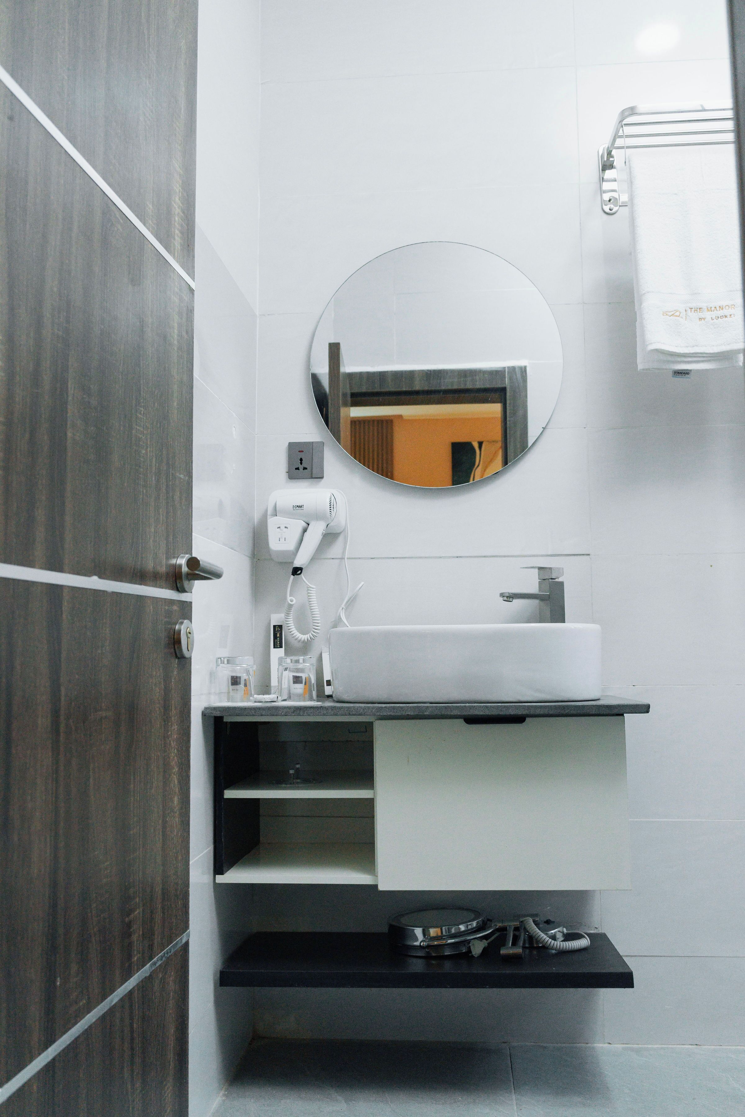 Un baño moderno con puerta de madera, un lavabo blanco, un espejo redondo y un perchero para toallas.