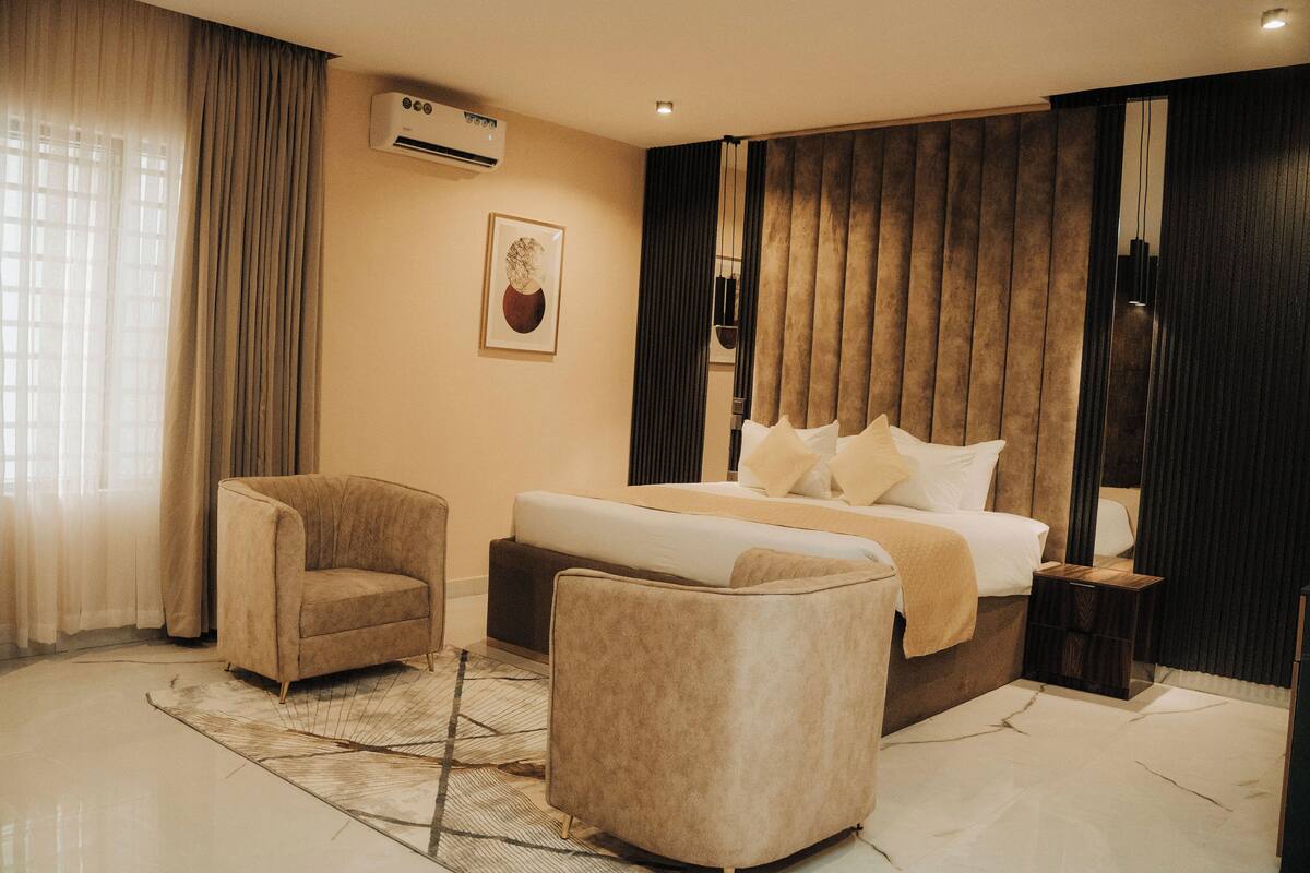 A modern hotel room with a large bed, two armchairs, a small side table, and a decorative wall piece.