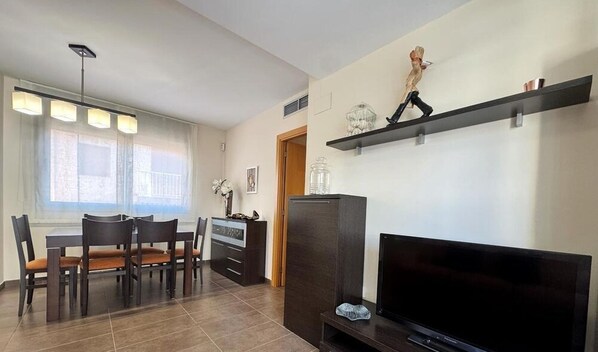 Una sala de estar moderna con un televisor de pantalla plana, una zona de comedor con mesa y sillas, y un estante decorativo con objetos.