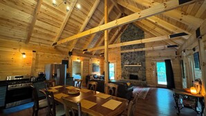 Une cabane spacieuse avec un plafond haut, des poutres en bois, une cheminée en pierre, un coin repas et une cuisine.