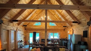Un intérieur spacieux de chalet en bois, avec un plafond haut, un coin salon central comprenant un canapé et un pouf, une table à manger et un coin cuisine.