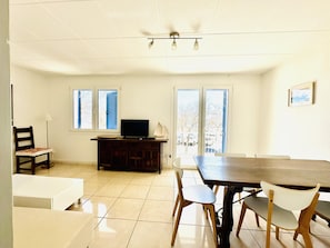 A dining area with a table and chairs, a television on a cabinet, and windows with a view of the outside.