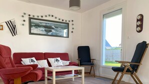 Een woonkamer met een rode bank, een witte salontafel, een blauwe fauteuil en een houten klapstoel. Een groot schilderij van een zeelandschap hangt aan de muur.