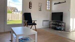 Een woonkamer met een witte tafel, een blauwe stoel, een tv op een meubel en een raam met uitzicht op een achtertuin.